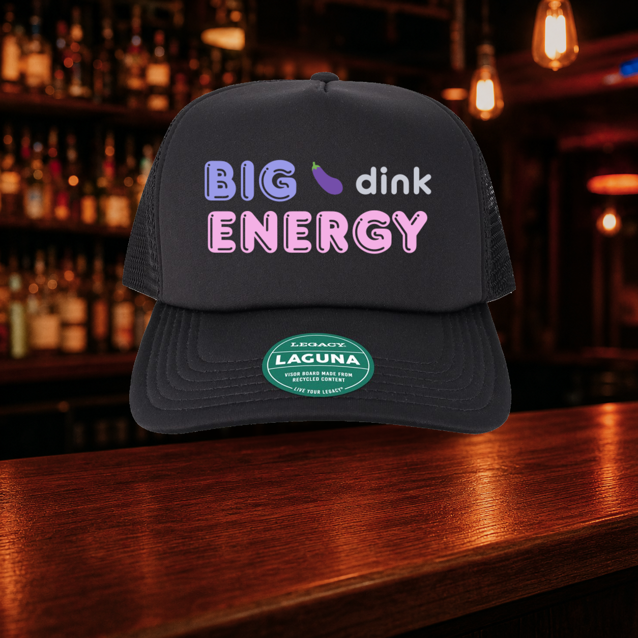 Big Dink Energy Trucker Hat shown against a warm after-dark bar backdrop