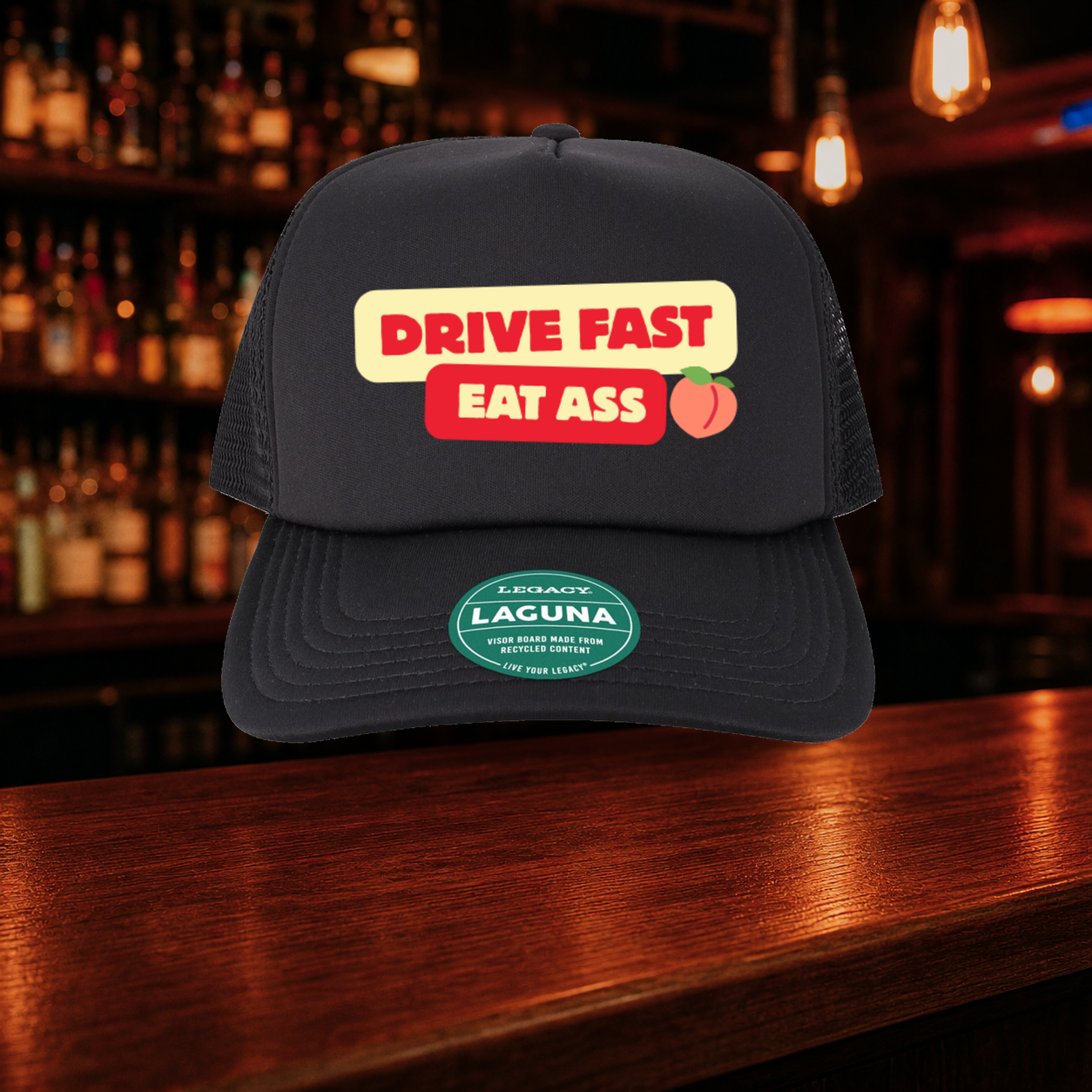 Eat A$$ Trucker Hat shown against a warm after-dark bar backdrop