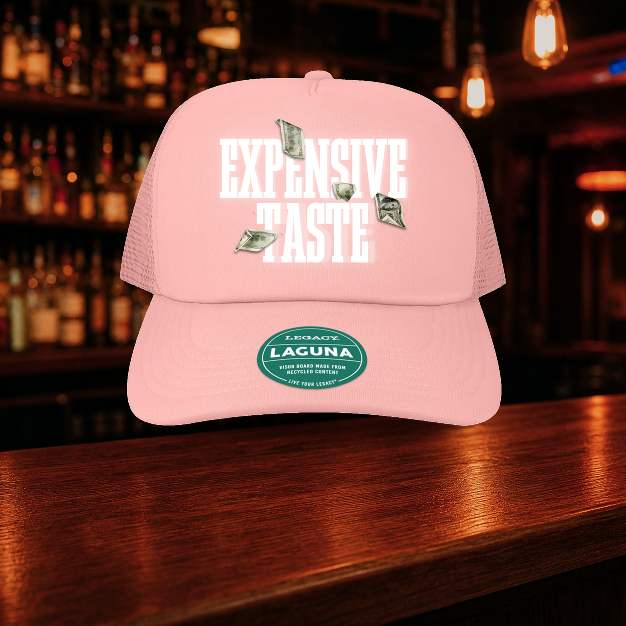 Expensive Taste Trucker Hat — Pink variety shown against a warm after-dark bar backdrop