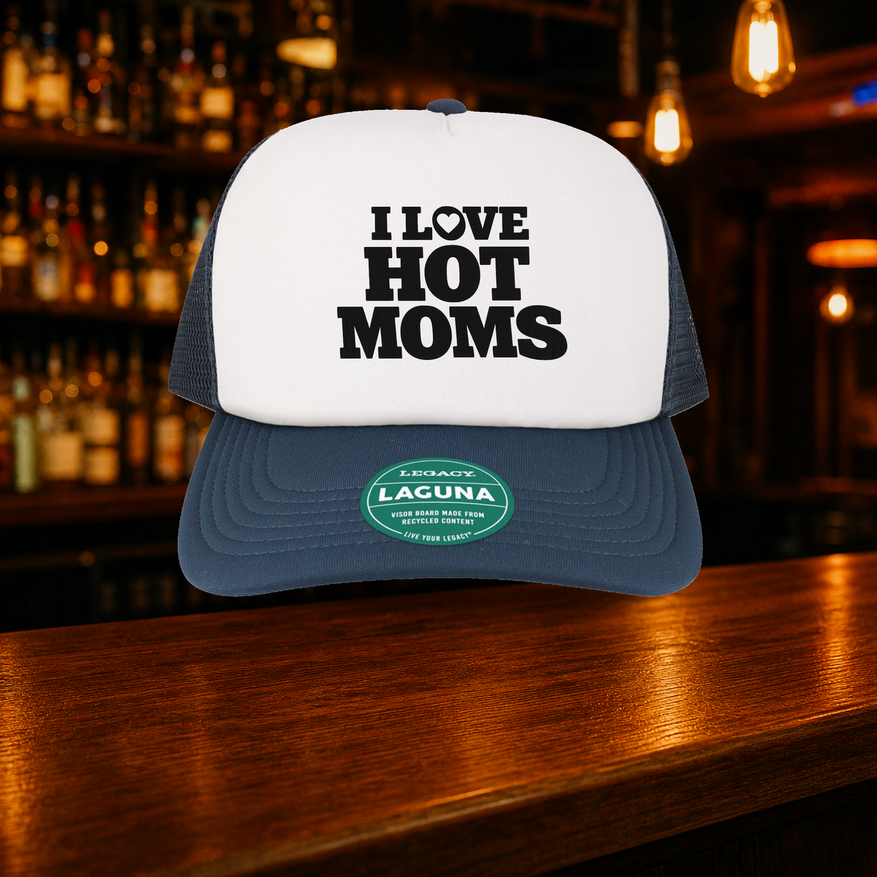 I Love Hot Moms Trucker Hat shown against a warm after-dark bar backdrop