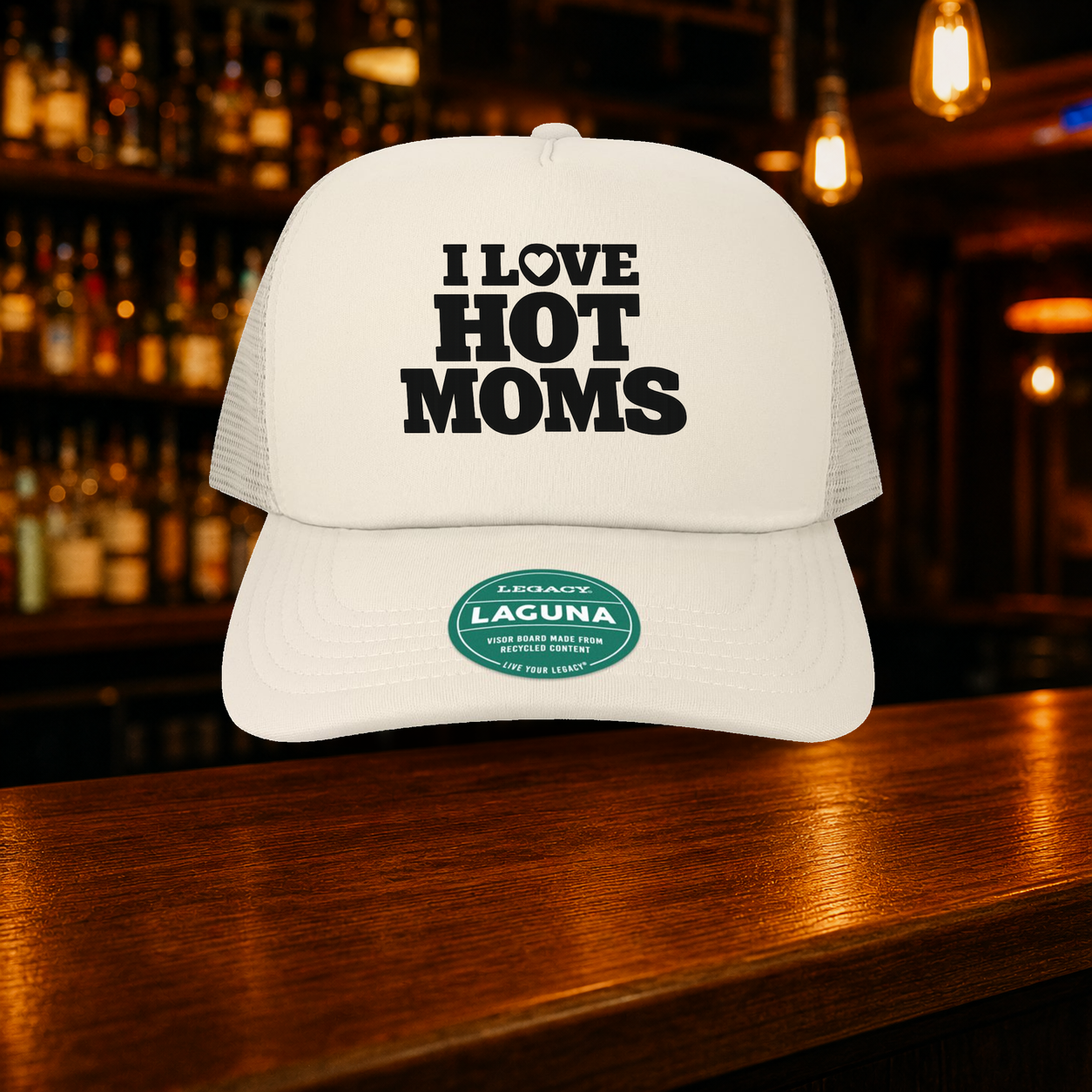 I Love Hot Moms Trucker Hat — Beige variety shown against a warm after-dark bar backdrop