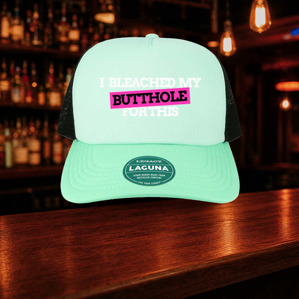 I Bleached My Butthole for This Trucker Hat — Teal variety shown against a warm after-dark bar backdrop