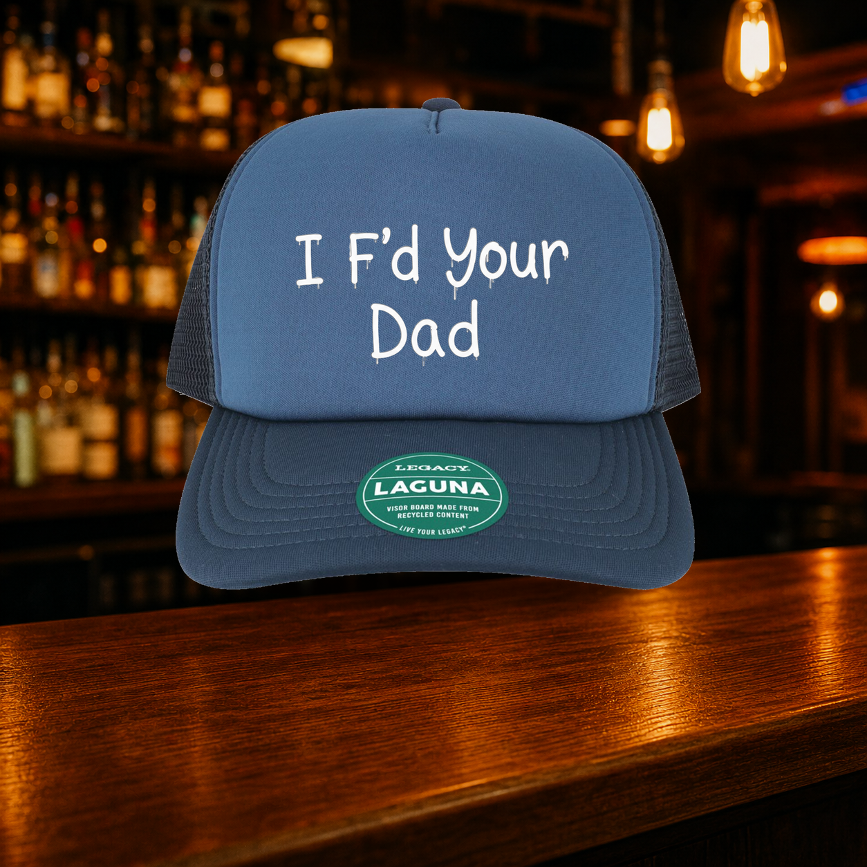 I F'd Your Dad Trucker Hat — Blue variety shown against a warm after-dark bar backdrop