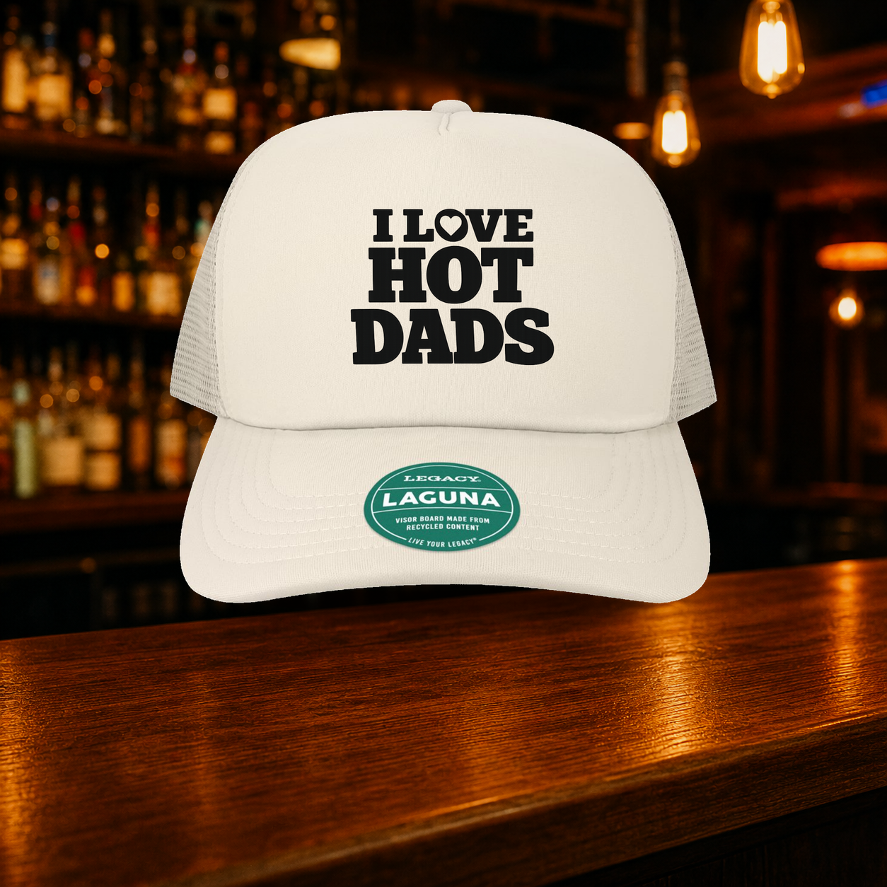 I Love Hot Dads Trucker Hat — Beige variety shown against a warm after-dark bar backdrop