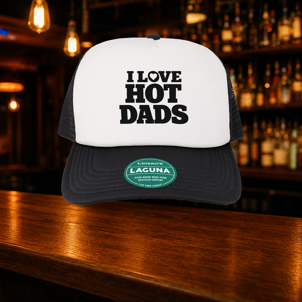 I Love Hot Dads Trucker Hat — Black variety shown against a warm after-dark bar backdrop