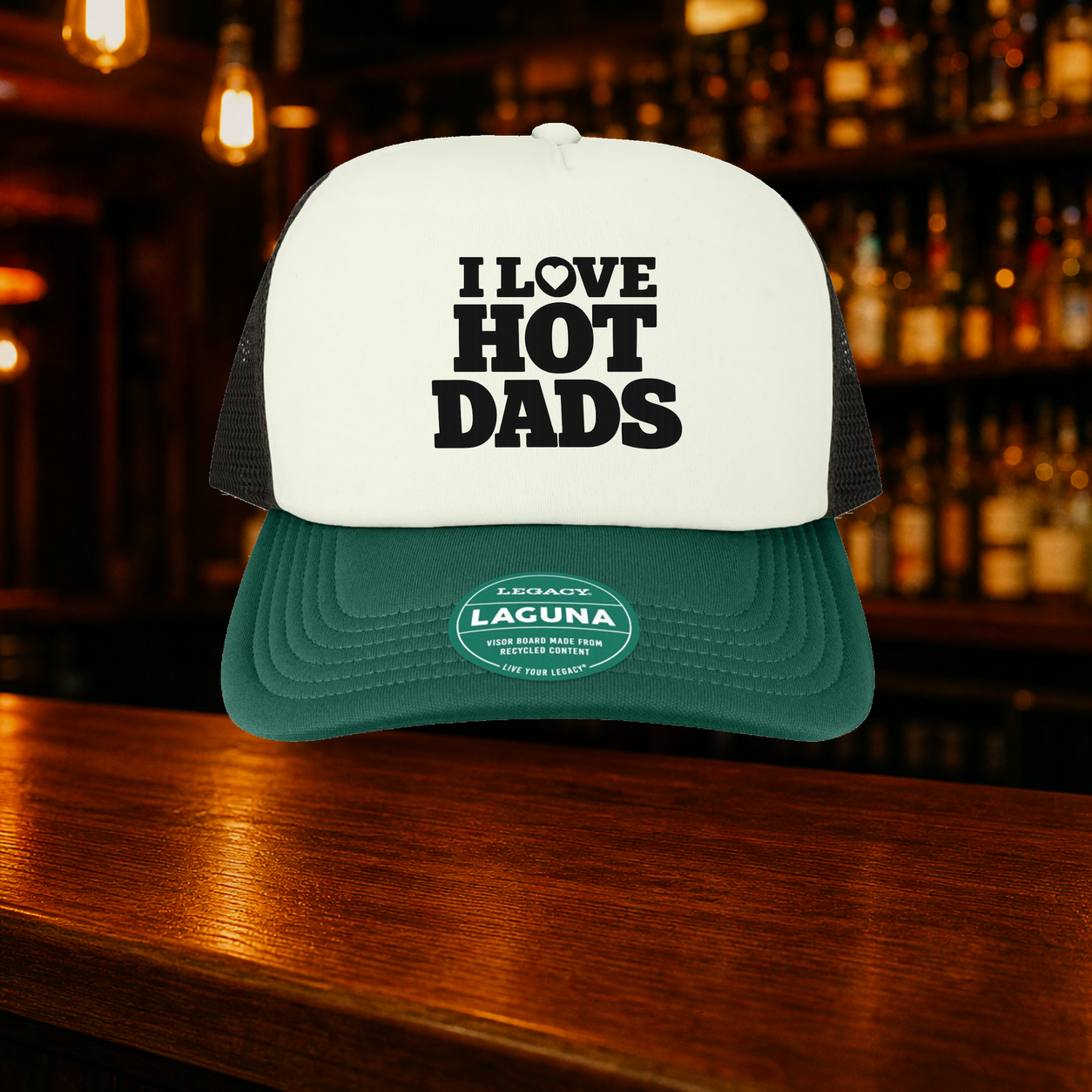 I Love Hot Dads Trucker Hat — Green variety shown against a warm after-dark bar backdrop