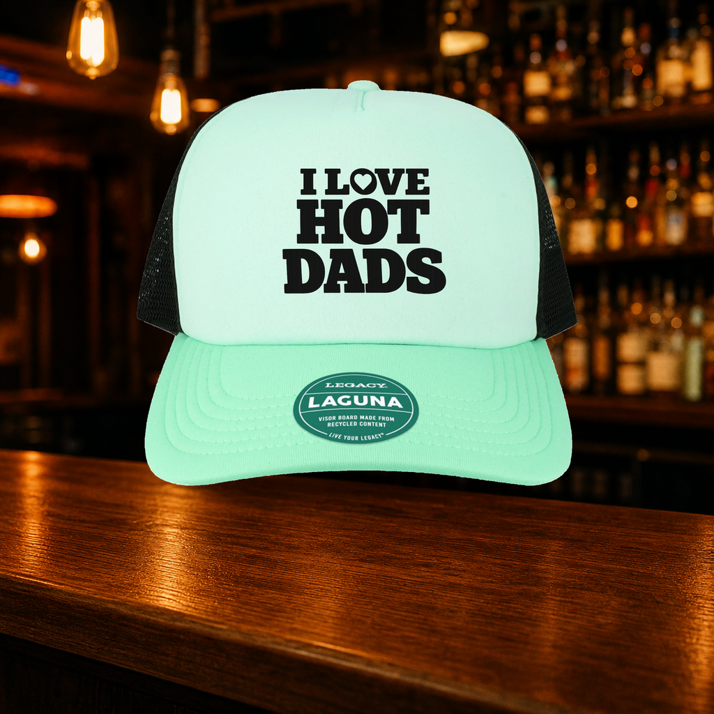 I Love Hot Dads Trucker Hat — Teal variety shown against a warm after-dark bar backdrop