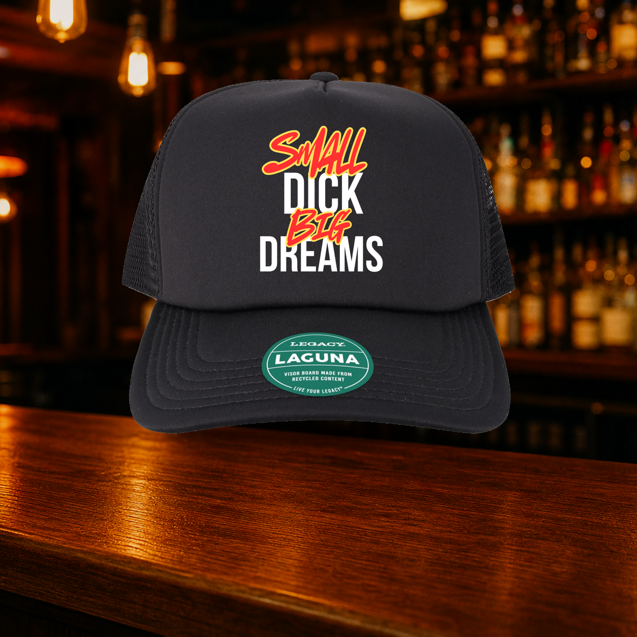 Small Dick Big Dreams Trucker Hat shown against a warm after-dark bar backdrop