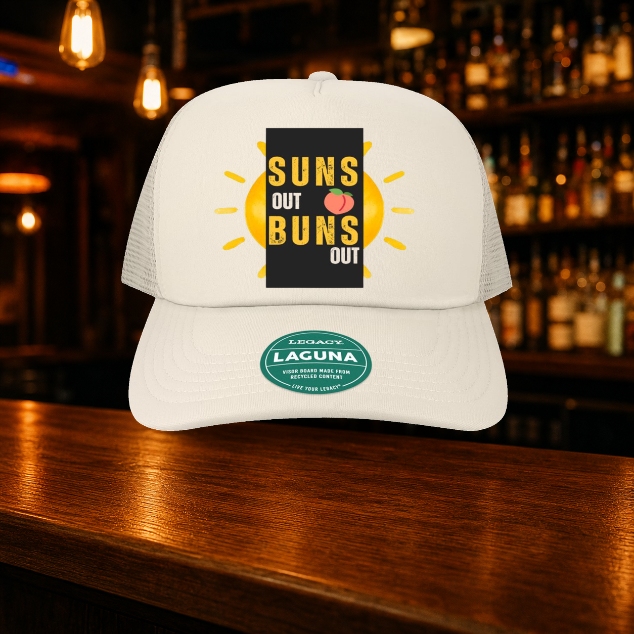 Suns Out Buns Out Trucker Hat — Beige variety shown against a warm after-dark bar backdrop