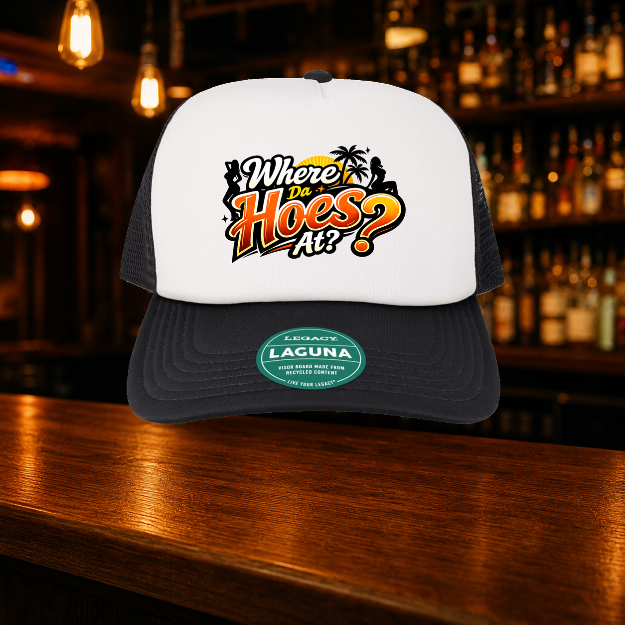 Where Da Hoes At? Trucker Hat — Black variety shown against a warm after-dark bar backdrop